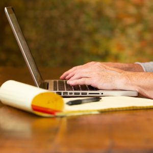 man typing on a computer laptop keyboard, legal pad with pen sitting on the wood table