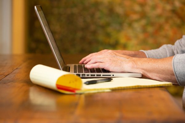 man typing on a computer laptop keyboard, legal pad with pen sitting on the wood table