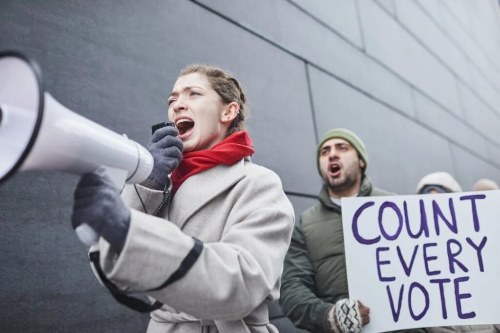 Image of a woman yelling into a loudspeaker and a man holding up a count every vote sign behind her. 