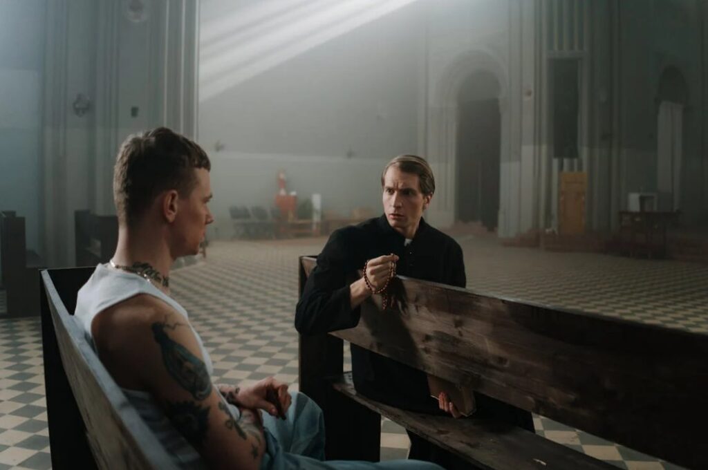 Image of two men talking in a church, one covered in tattoos, and one a priest. 