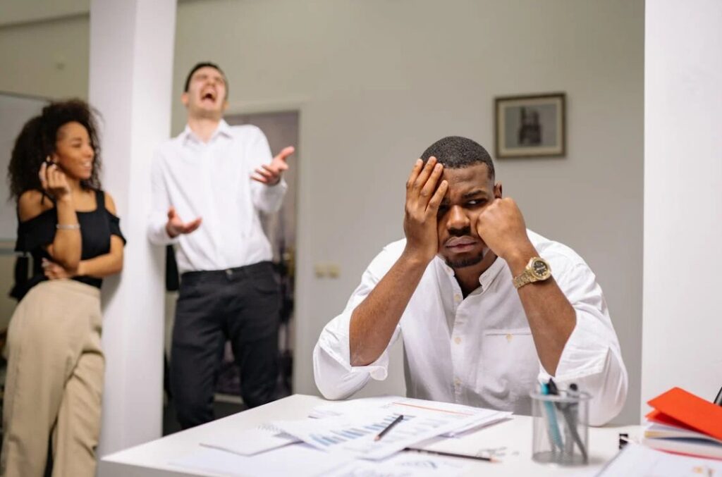 Image of a frustrated man sitting with a man and woman laughing nearby him. 