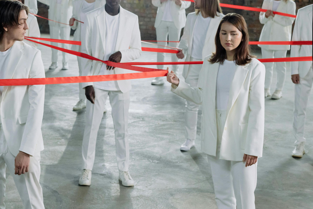 A woman stands apart from a crowd, holding the same red line of fate that everyone else does, tethered to the rest, but her mind drifts elsewhere. A man in the corner of the frame looks to her with a sort of intrigue