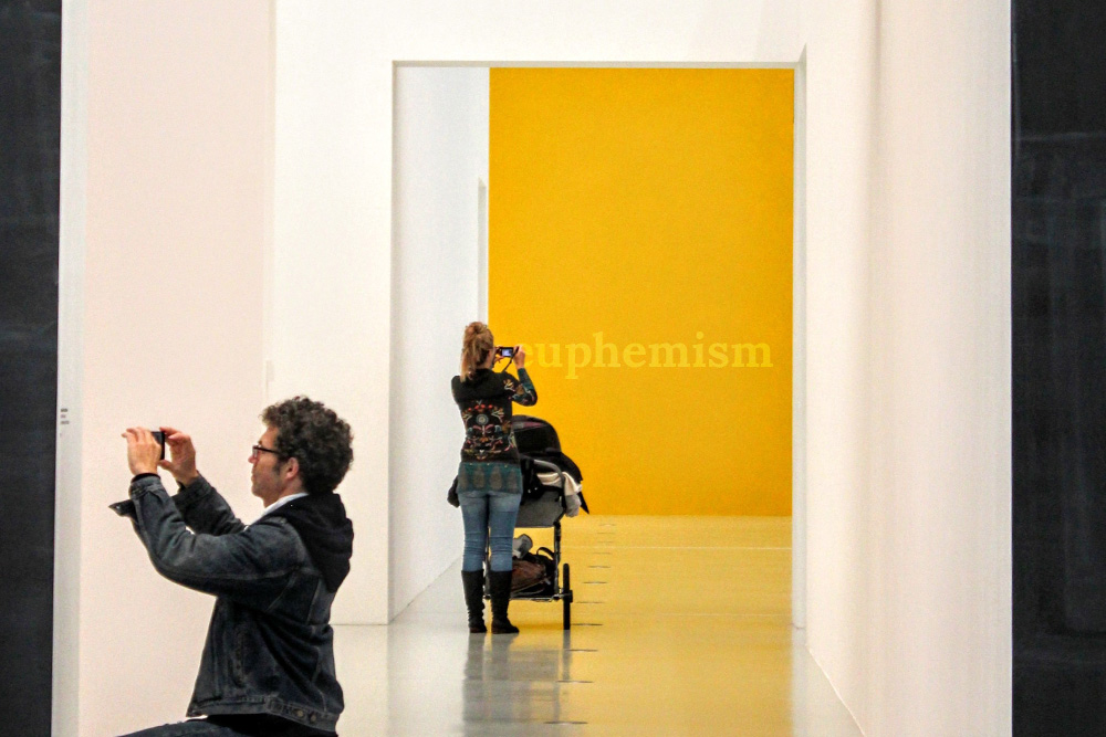 People in an art gallery taking photos of different walls, a woman with a child in a carriage taking a picture of a wall with the word "Euphemism" on it to visualize the art of euphemism