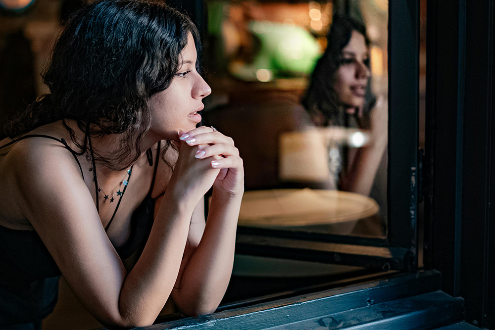A woman who is pensive and furtive, and her reflection beside her smiling, suggesting the dichotomy of an internal dialogue as perceived by a viewer