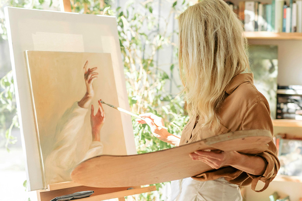 A nondescript white woman paints a realistic image of hands for the blog realistic fiction. perhaps the writer reading the article may be able to imagine themselves as a painter making something realistic too