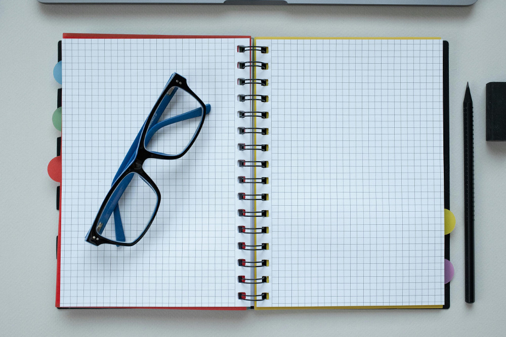 An open graph paper notebook with a pair of glasses set to one page and a laptop looming just a little bit at the top of the image nearby. A coldness of the empty page suggested by the blue coloring