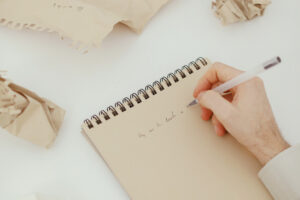 A writer is trying to rewrite a simple thing on a notepad with crumpled pieces of paper all around. Beauty and lightness are evoked in the frame, suggesting this as a necessary process.