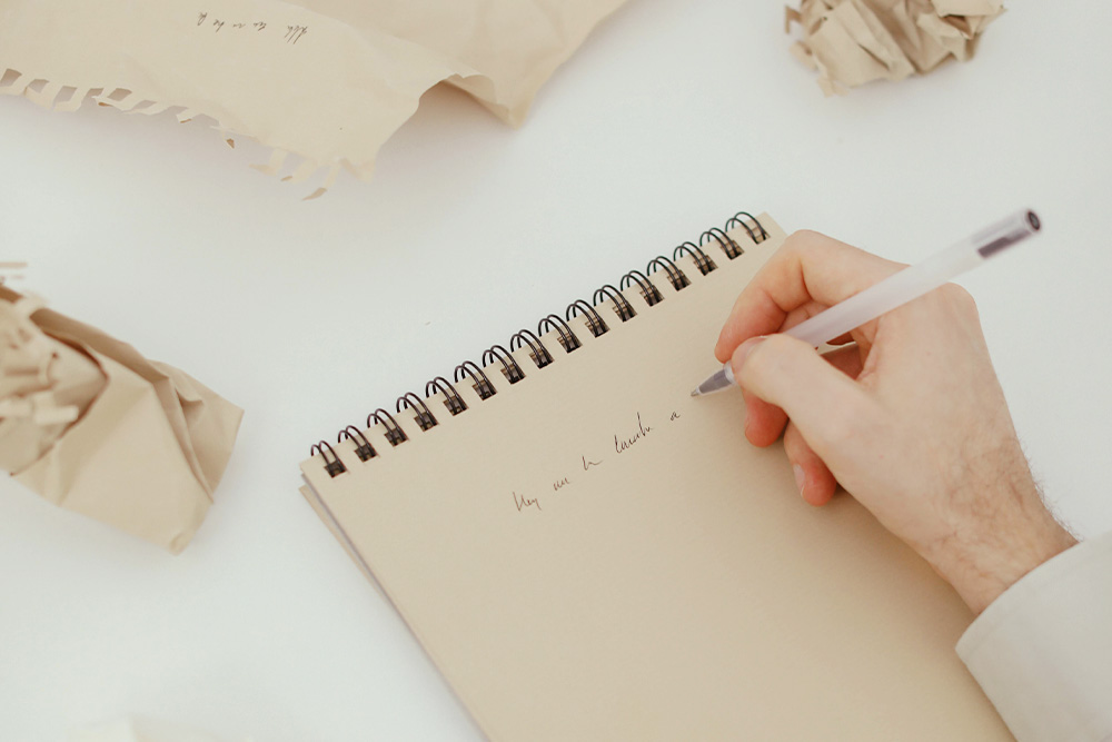 A writer is trying to rewrite a simple thing on a notepad with crumpled pieces of paper all around. Beauty and lightness are evoked in the frame, suggesting this as a necessary process.