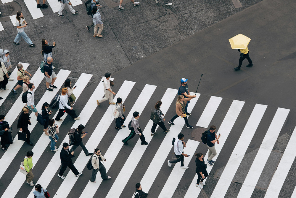 Someone in an incognito getup under a yellow umbrella crosses against the grain of a crowd at an intersection to suggest external conflict in the context of character vs. society.