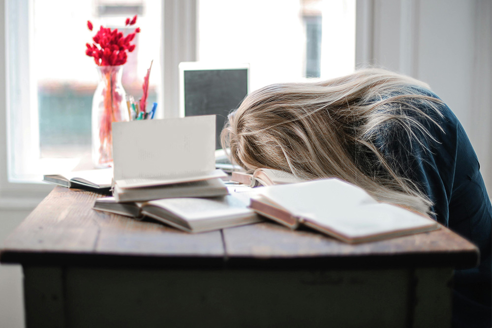 A woman with her head sunk into a pile of books is a visual simile for the author reading this article to learn overcoming procrastination