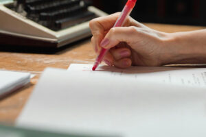 A hand writing on papers with a red pen suggests the romance that can be found in the process of proofreading work