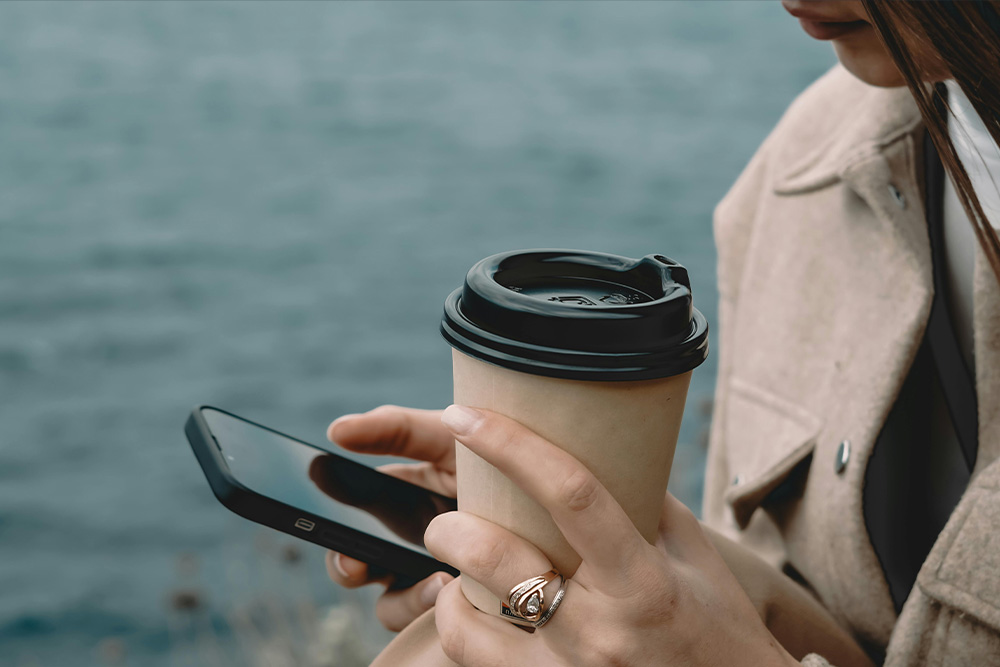 An image of a woman smiling while scrolling on her phone with a coffee in hand suggesting austerity, enjoyment, peace from looking at writing memes