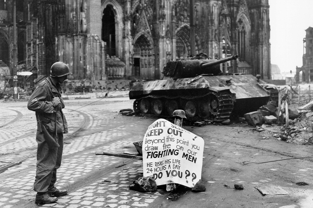A sullen soldier stands at the side of a sign and tank to suggest that he will pursue war inevitably at is his imperative, and that every soldier fights a different battle. Struggle of the protagonist.