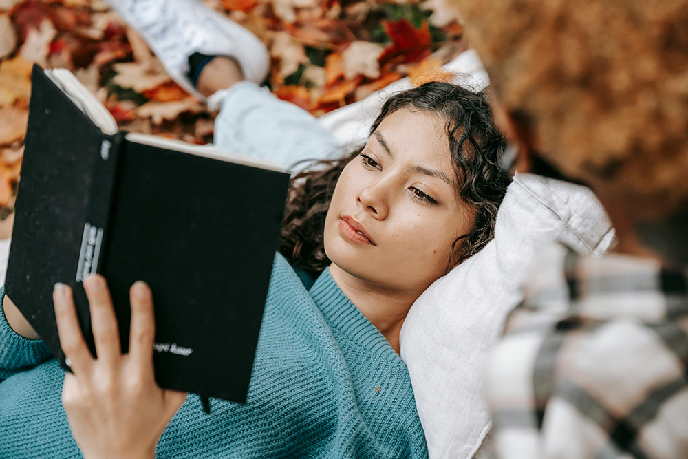 A woman reading a book wistfully in fall foliage to represent Memoir writing prompts