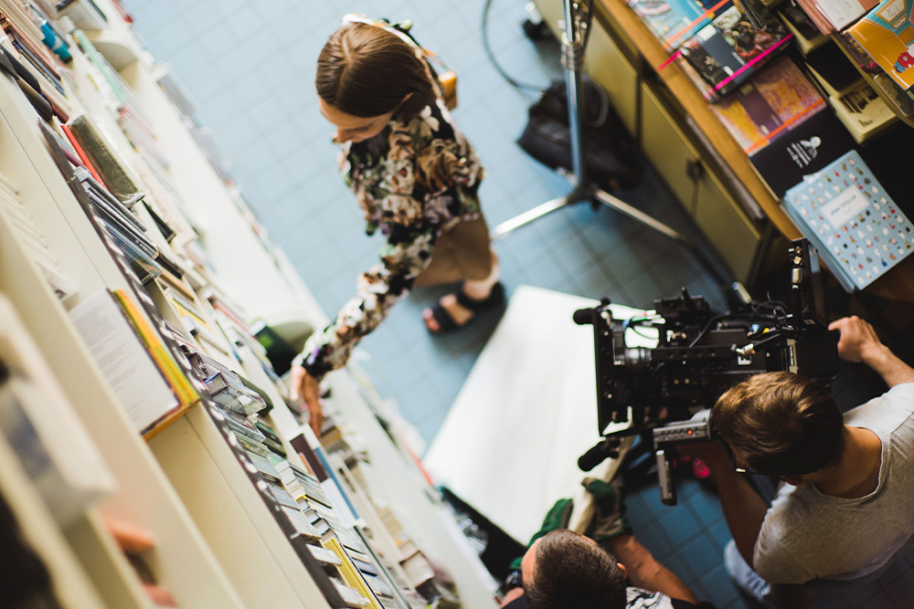A woman in a bookshop look at novels with a camera beside her as a visual parallel for how to adapt a book into a screenplay