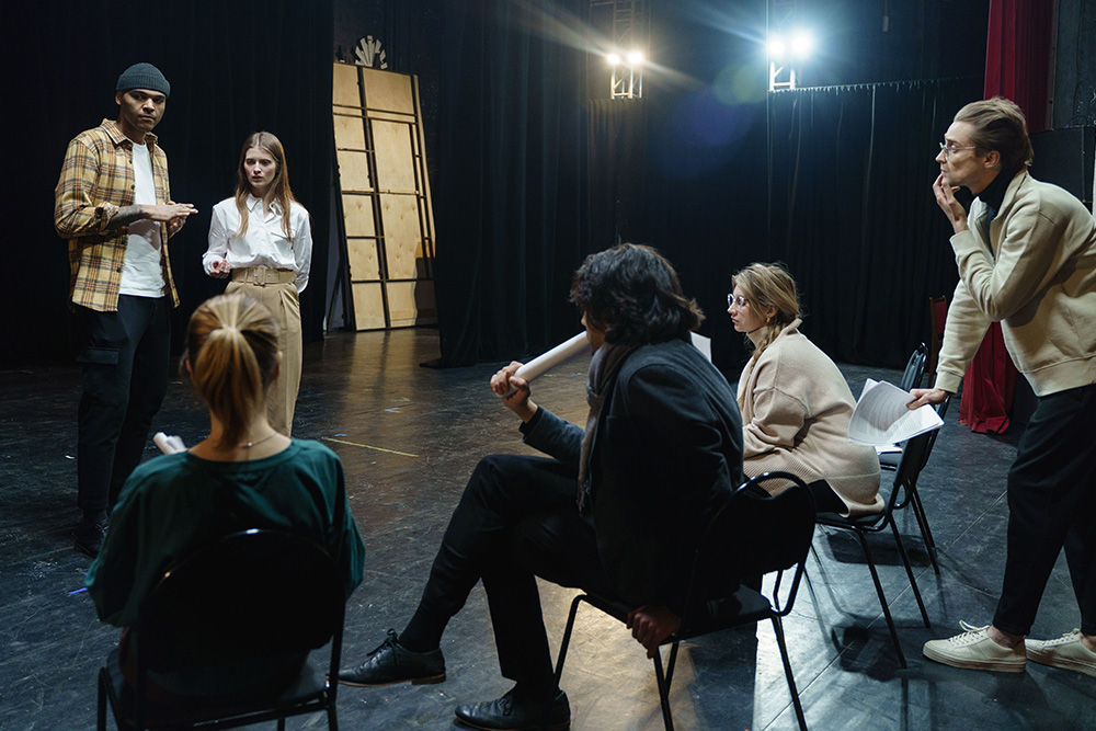 A group of actors and writers and a director sit with pages in hand around two actors under the glow of light — how many words a scene needs must be visualized as you pretend to be the director and actor to choose the effective amount when asking how many words per page in a book?