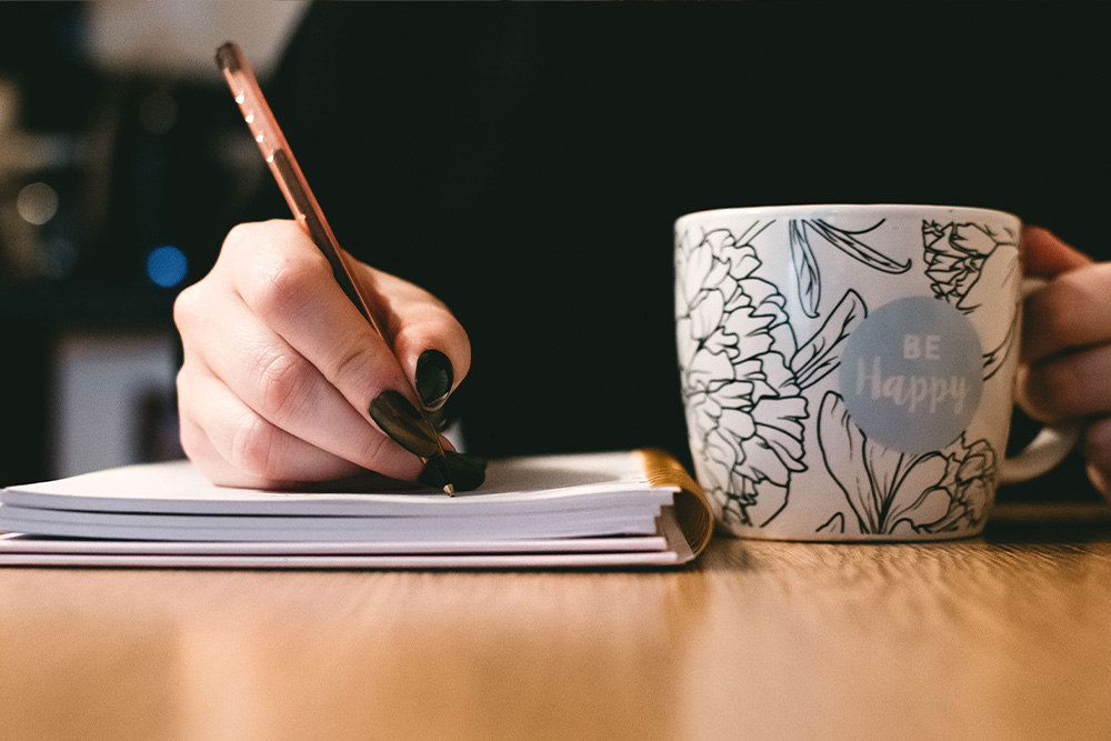 Peaceful picture of a woman writing in a journal with a coffee cup beside her to suggest how to write an author bio