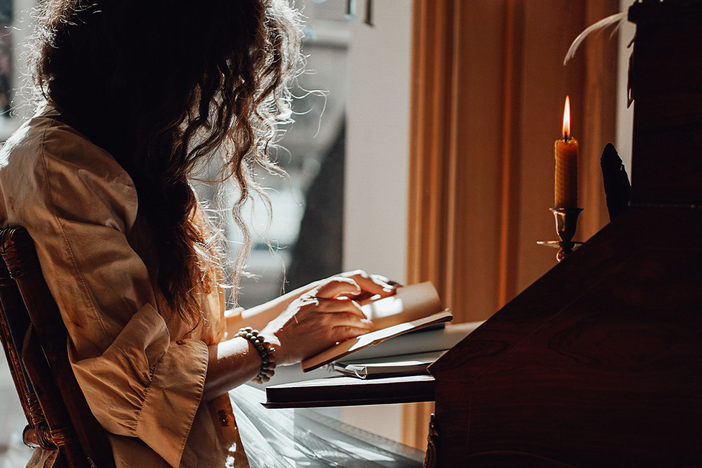 A woman at a writerly table with a sunbeam and candles adorning her workspace