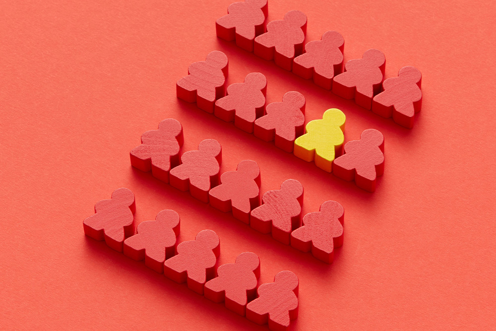 A little yellow wooden person block stands out in color against crowd of red wooden people